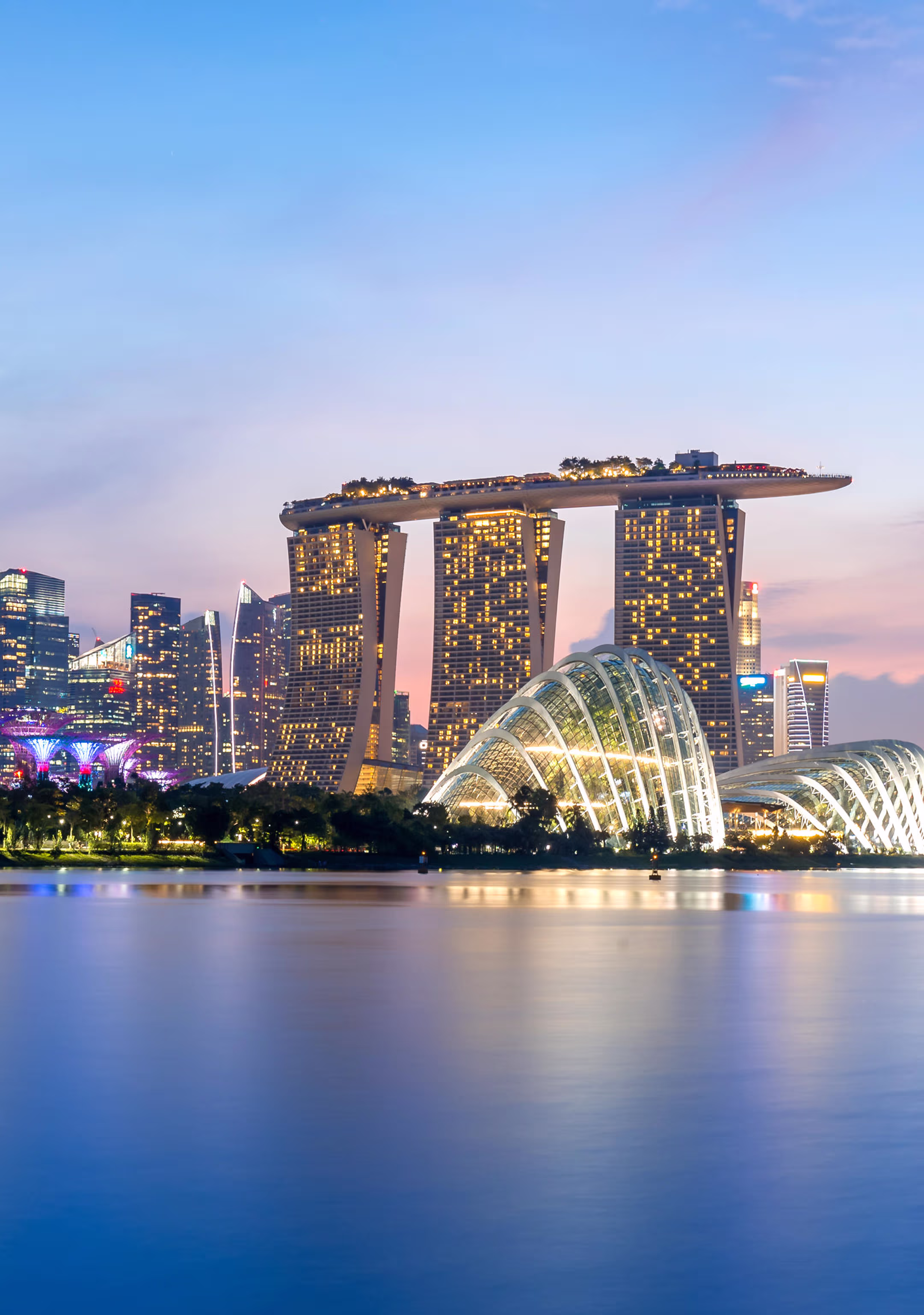 Singapore skyline at dusk featuring Marina Bay Sands hotel and illuminated glass domes of Gardens by the Bay reflecting on calm water.