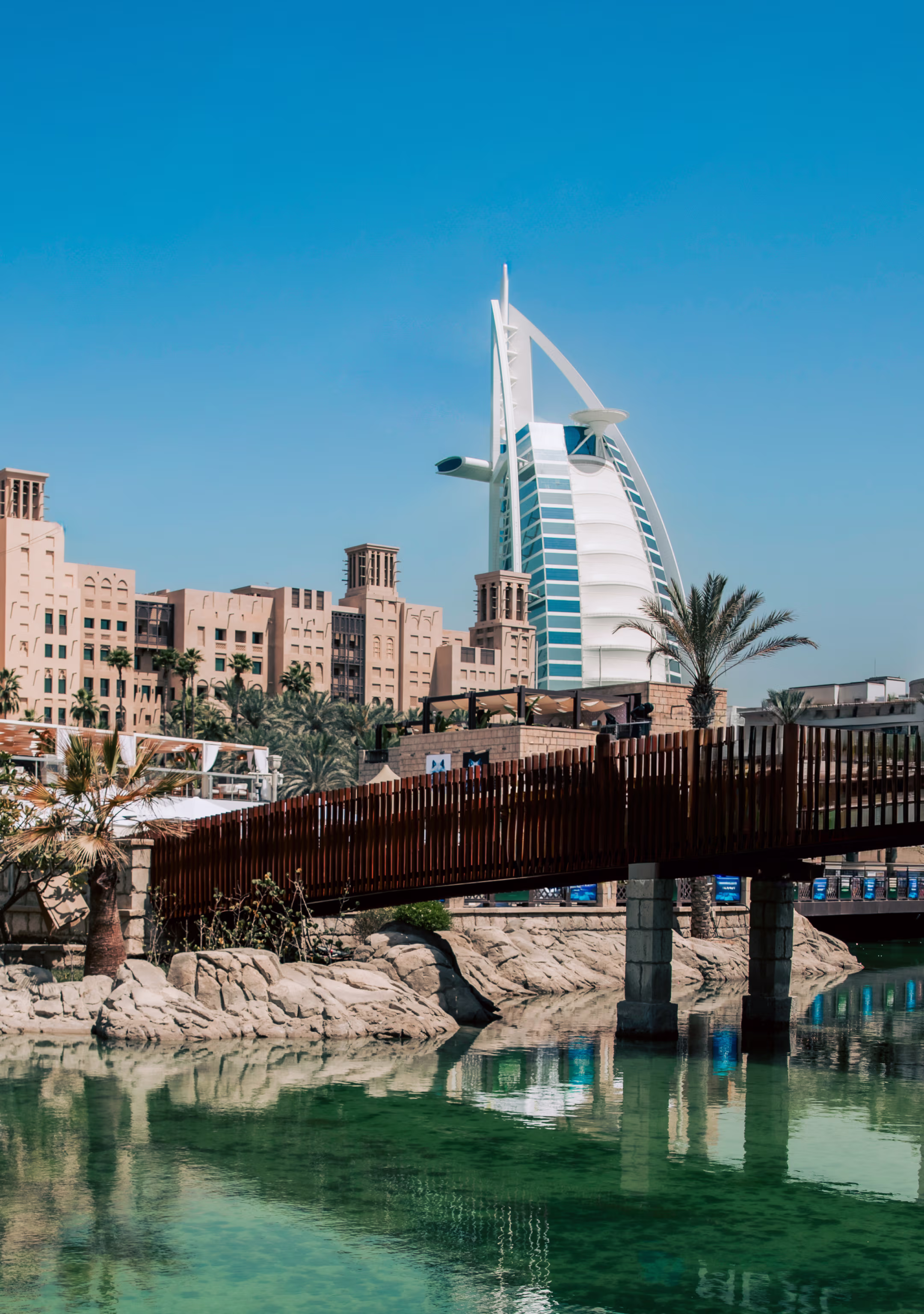 Burj Al Arab hotel with traditional buildings, palm trees, a wooden bridge, and clear water reflecting the scene in Dubai.