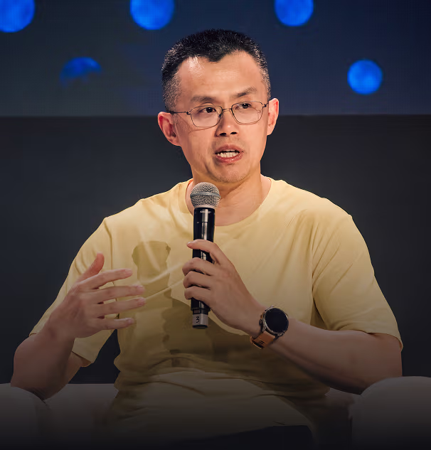 Man wearing glasses and a yellow t-shirt speaking into a microphone with a dark blurred background.