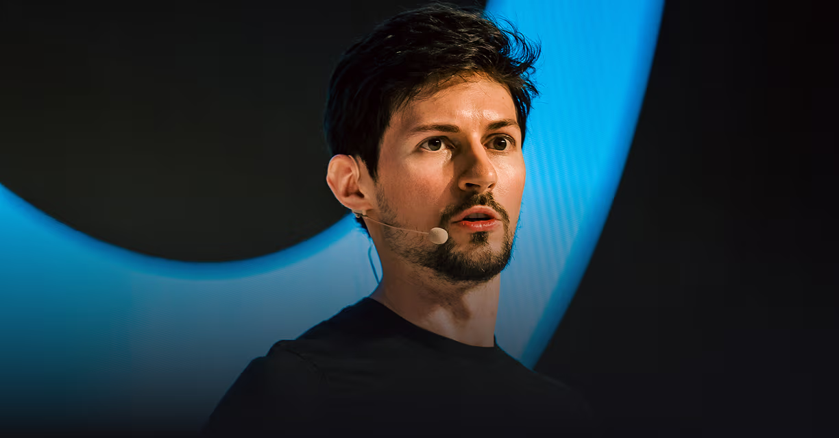 Young man with a beard wearing a headset microphone, speaking in front of a large blue curved light on a dark background.